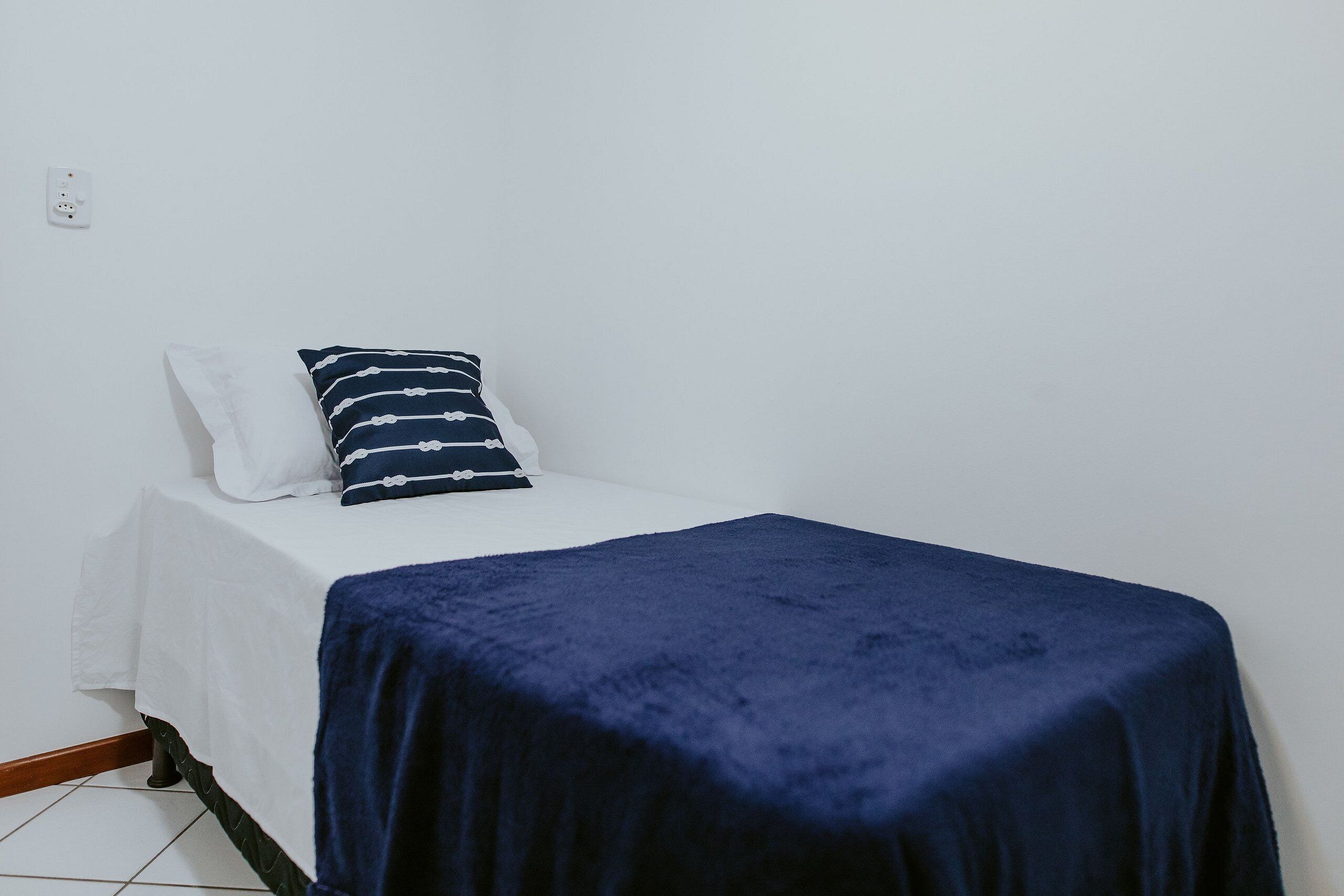 A single bed with a white mattress and a dark blue blanket, a white pillow, and a striped pillow on a white wall with a power outlet.