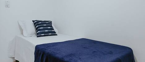 A single bed with a white mattress and a dark blue blanket, a white pillow, and a striped pillow on a white wall with a power outlet.