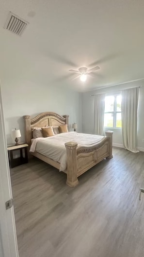 Ein Schlafzimmer mit einem großen Bett, einem Deckenventilator, einem Nachttisch mit Lampe und einem Fenster mit Vorhängen.