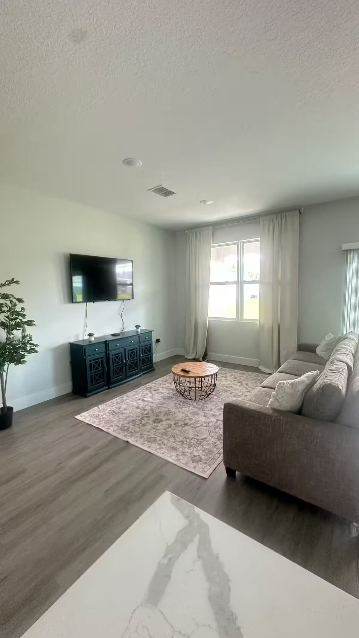 Un salon moderne avec un téléviseur à écran plat, un meuble de rangement pour appareils électroniques de couleur foncée, une table basse ronde, un canapé beige et une grande fenêtre avec des rideaux.