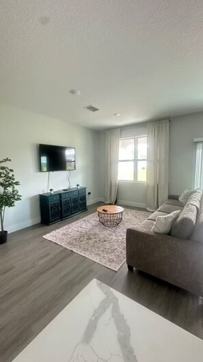 A modern living room with a flat-screen TV, a dark-colored entertainment unit, a round coffee table, a beige sofa, and a large window with curtains. - The Grey House (Palm Bay)