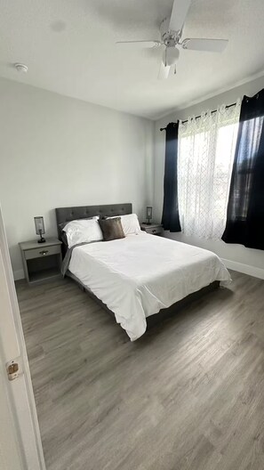 A bedroom with a bed, nightstands, a ceiling fan, and a window with curtains. - The Grey House (Palm Bay)