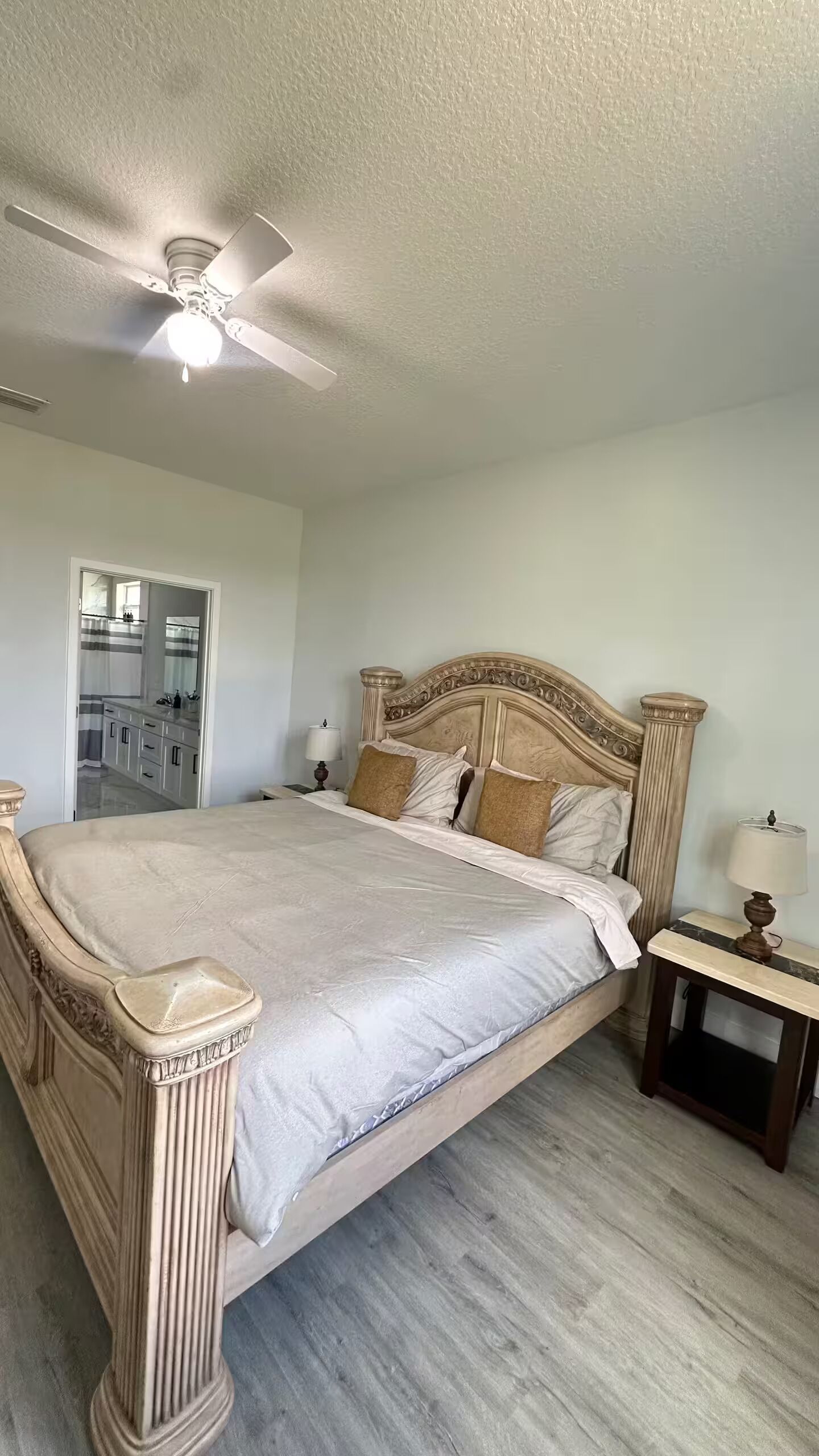 A bedroom with a large bed, a ceiling fan, a nightstand, and a bathroom visible through an open door.