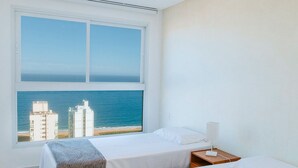 A hotel room with two beds, a large window with a view of the ocean and buildings, and a nightstand with a lamp. - Modern apartment with incredible sea views (Vila Velha)