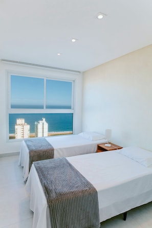Ein Hotelzimmer mit zwei Betten, einem großen Fenster mit Blick auf das Meer und Gebäude sowie einem Nachttisch mit einer Lampe.