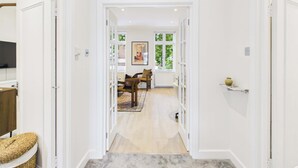 A hallway with white walls, a wooden floor, and a door leading to a room with a window and a painting. - Be London - The Hampstead Residences (Greater London)