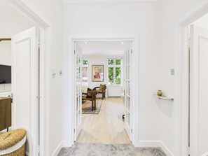 A hallway with white walls, a wooden floor, and a door leading to a room with a window and a painting. - Be London - The Hampstead Residences (Greater London)
