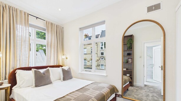 A bedroom with a bed, a bedside table, a mirror, and a window with a view of buildings. - Be London - The Hampstead Residences (Greater London)