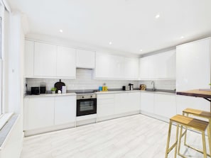 A modern kitchen with white cabinetry, a built-in oven, and a wooden bar stool. - Be London - The Hampstead Residences (Greater London)