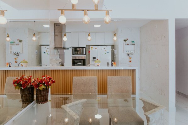 A modern kitchen with white cabinets, a central island, and a dining area with a glass table and chairs. - 3-Bedroom Spacious and Charming Apartment with Nature Views (Vila Velha)