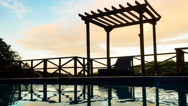 En pool med en pergola av trä, klar himmel och stilla vatten som speglar pergolan.