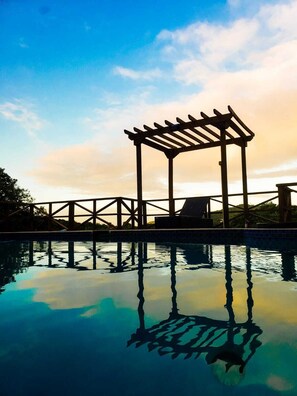 Una piscina con una estructura de pérgola de madera, cielo despejado y agua tranquila que refleja la estructura.