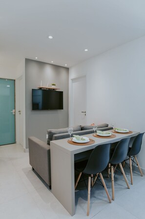 A modern dining area with a table set for four, a flat-screen TV mounted above, and a sofa in the background. - Modern 2-bedroom apartment with pool and gym. (Vila Velha)