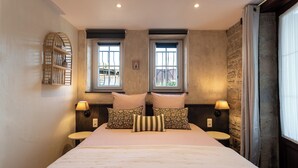 A hotel room with a large bed, two windows, a small table, and a wall-mounted shelf.