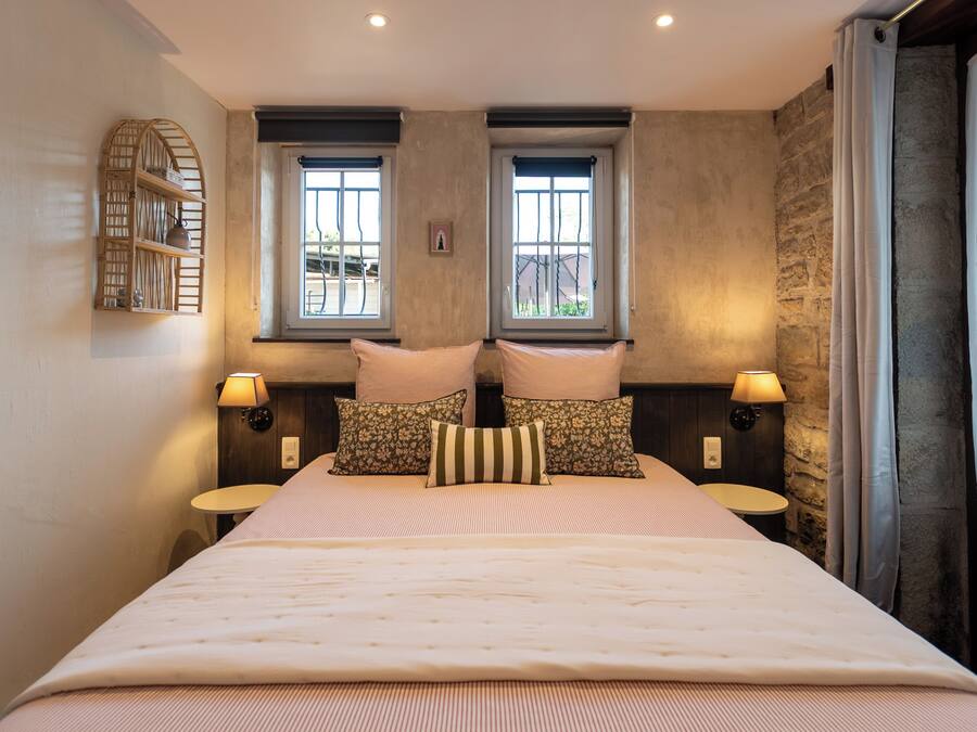 A hotel room with a large bed, two windows, a small table, and a wall-mounted shelf.