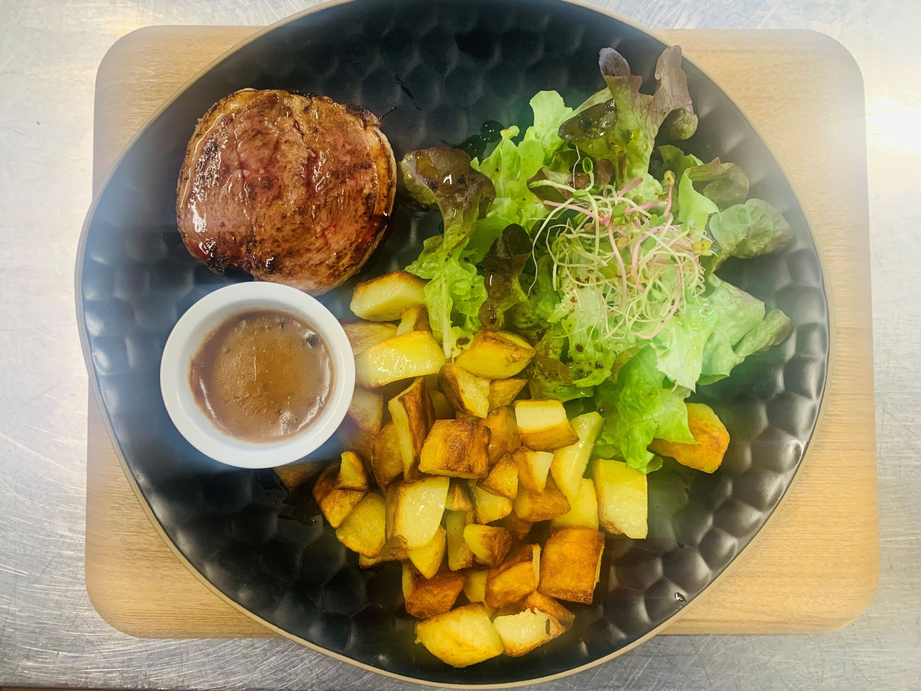 A plate with a grilled steak, roasted potatoes, a salad with greens and sprouts, and a small bowl of sauce.