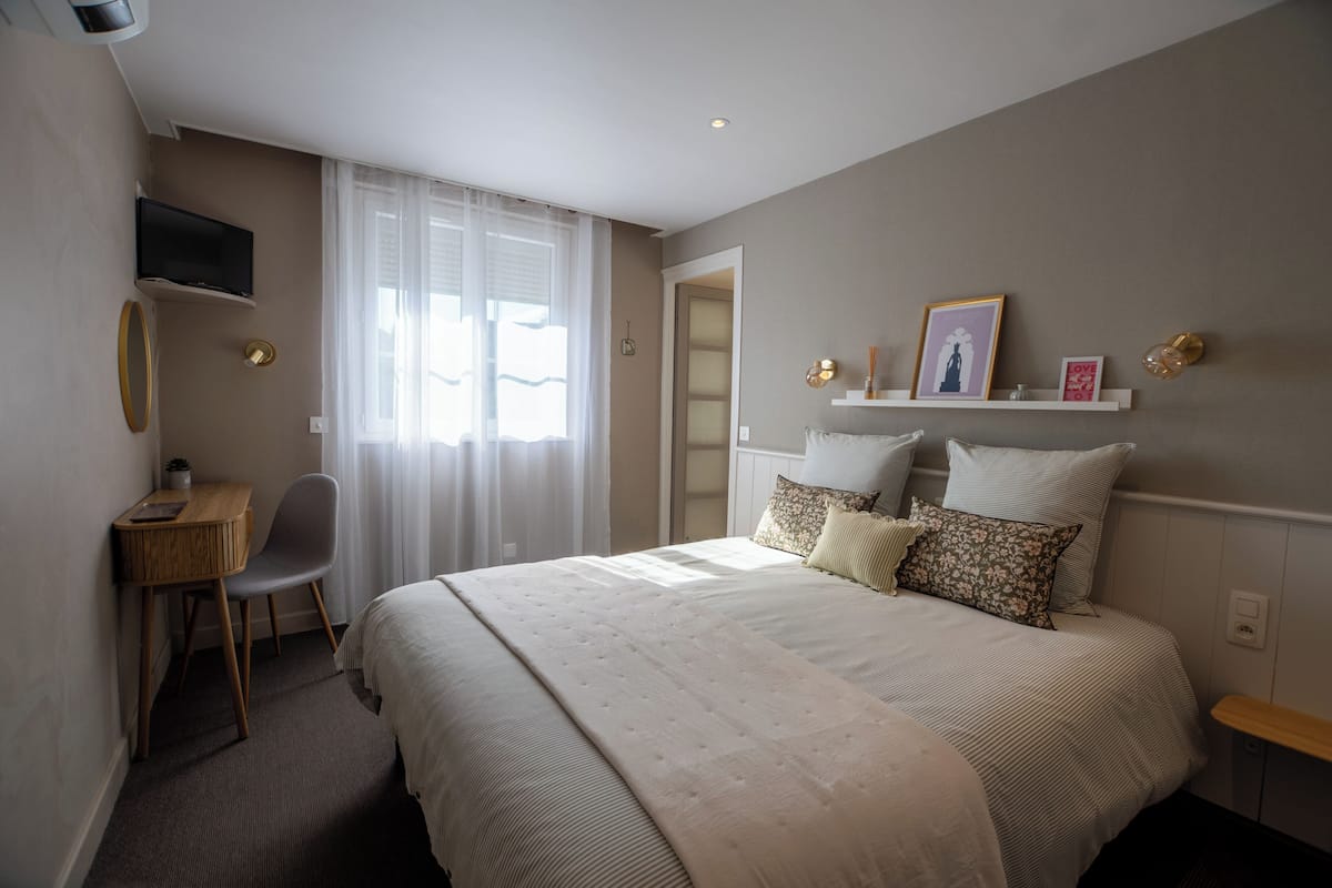A bedroom with a bed, a desk, a chair, a TV, and a window with curtains.