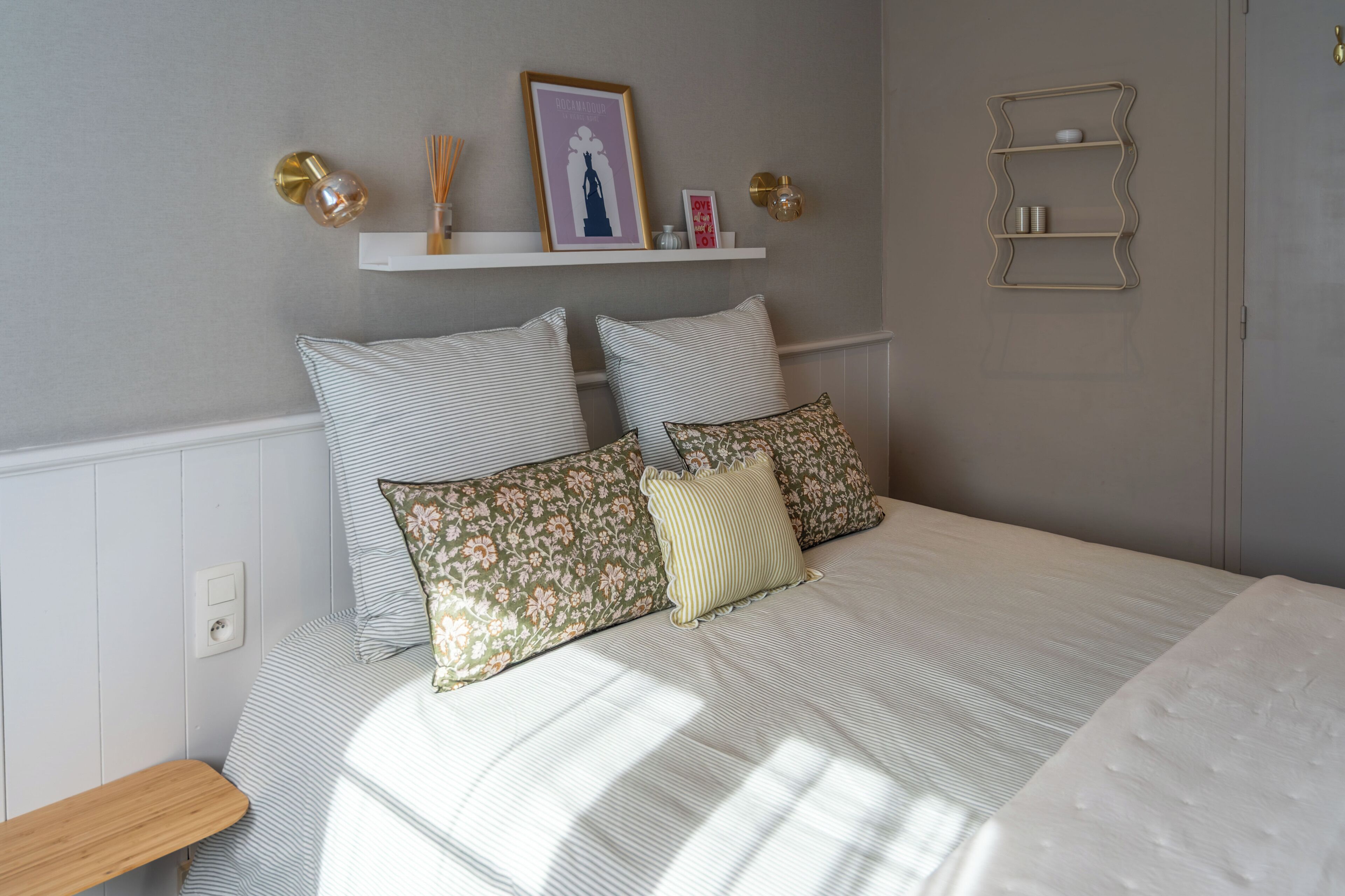 A neatly made bed with striped bedding, a wooden bedside table, and a wall-mounted shelf with decorative items.