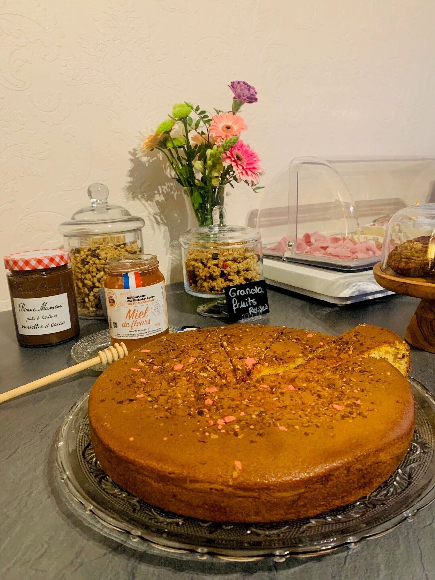 Ein Kuchen auf einer Glasplatte, Gläser mit Honig und Marmelade, eine Blumenvase und ein Schild für Granola-Früchte.
