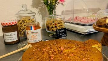 Ein Kuchen auf einer Glasplatte, Gläser mit Honig und Marmelade, eine Blumenvase und ein Schild für Granola-Früchte.