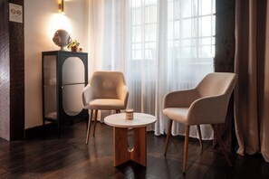 Una habitación con dos sillones, una mesita redonda, un aparador decorativo y un ventanal con cortinas.