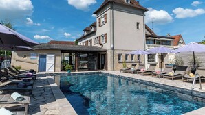 A swimming pool with lounge chairs and umbrellas, a building with a tiled roof, and a clear blue sky.