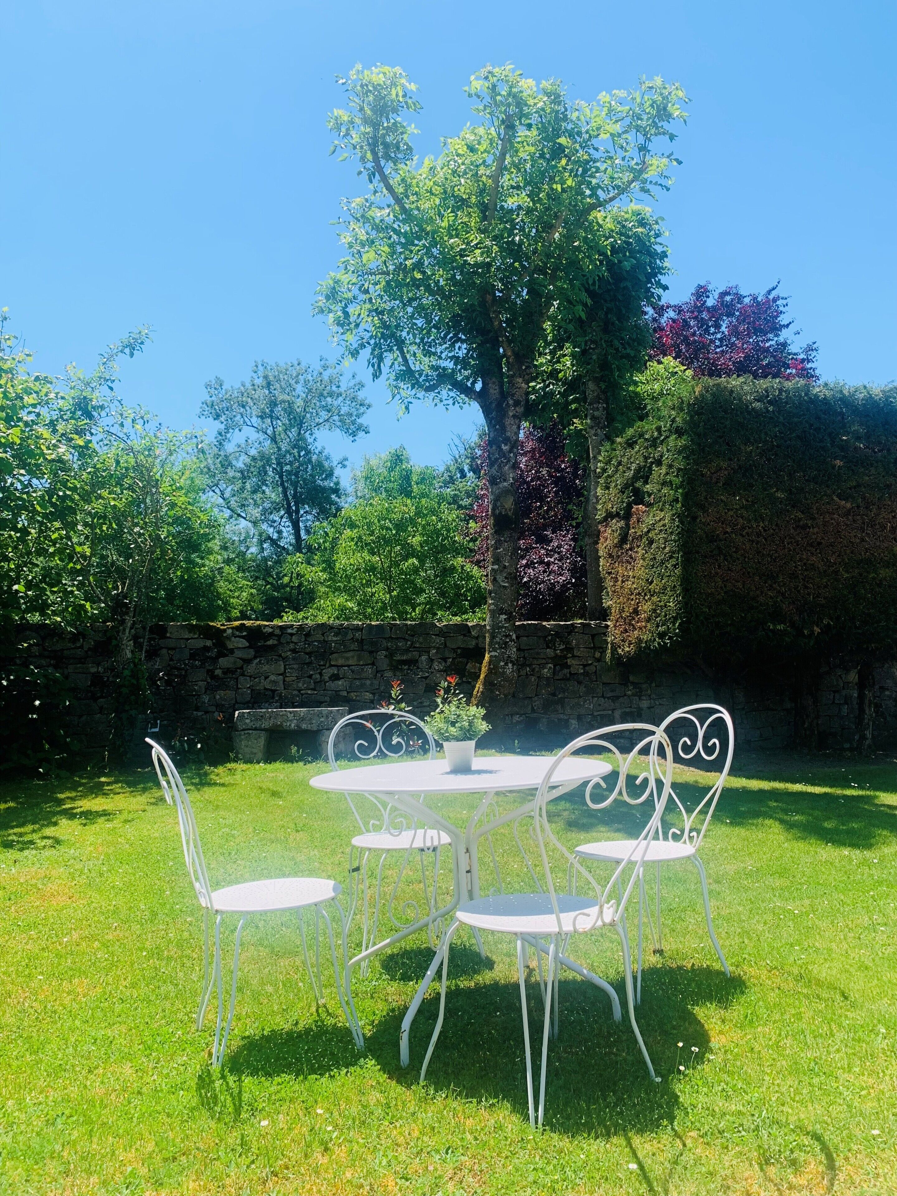 A white metal table and chairs set on a grassy lawn with a stone wall and trees in the background.