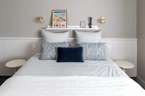 Une chambre à coucher bien rangée, avec un lit, deux tables de chevet, une étagère murale agrémentée d’objets décoratifs et un tableau encadré.