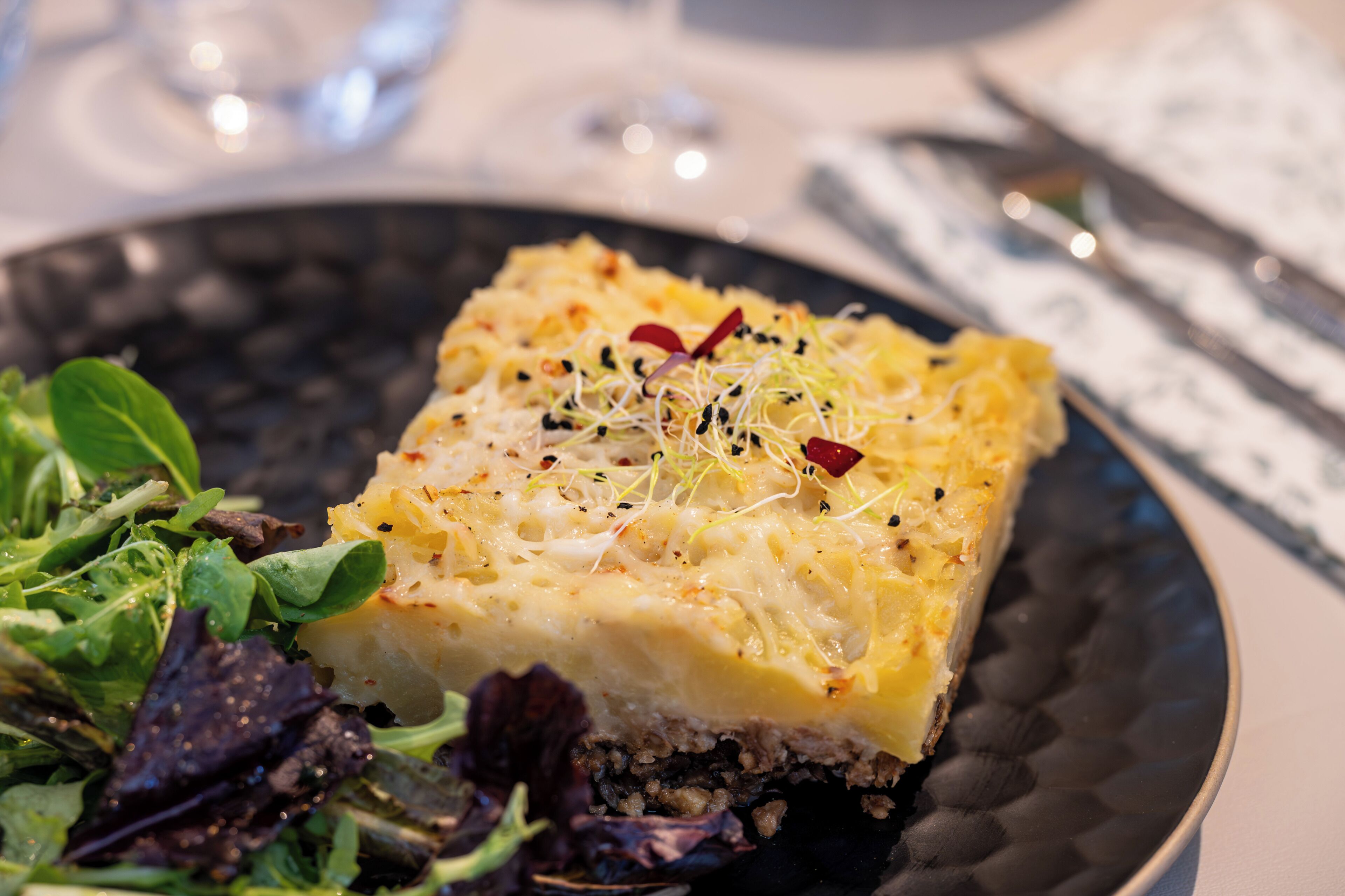 A square dish of food with a creamy top, garnished with microgreens and red pepper flakes, served on a dark plate with a side salad.