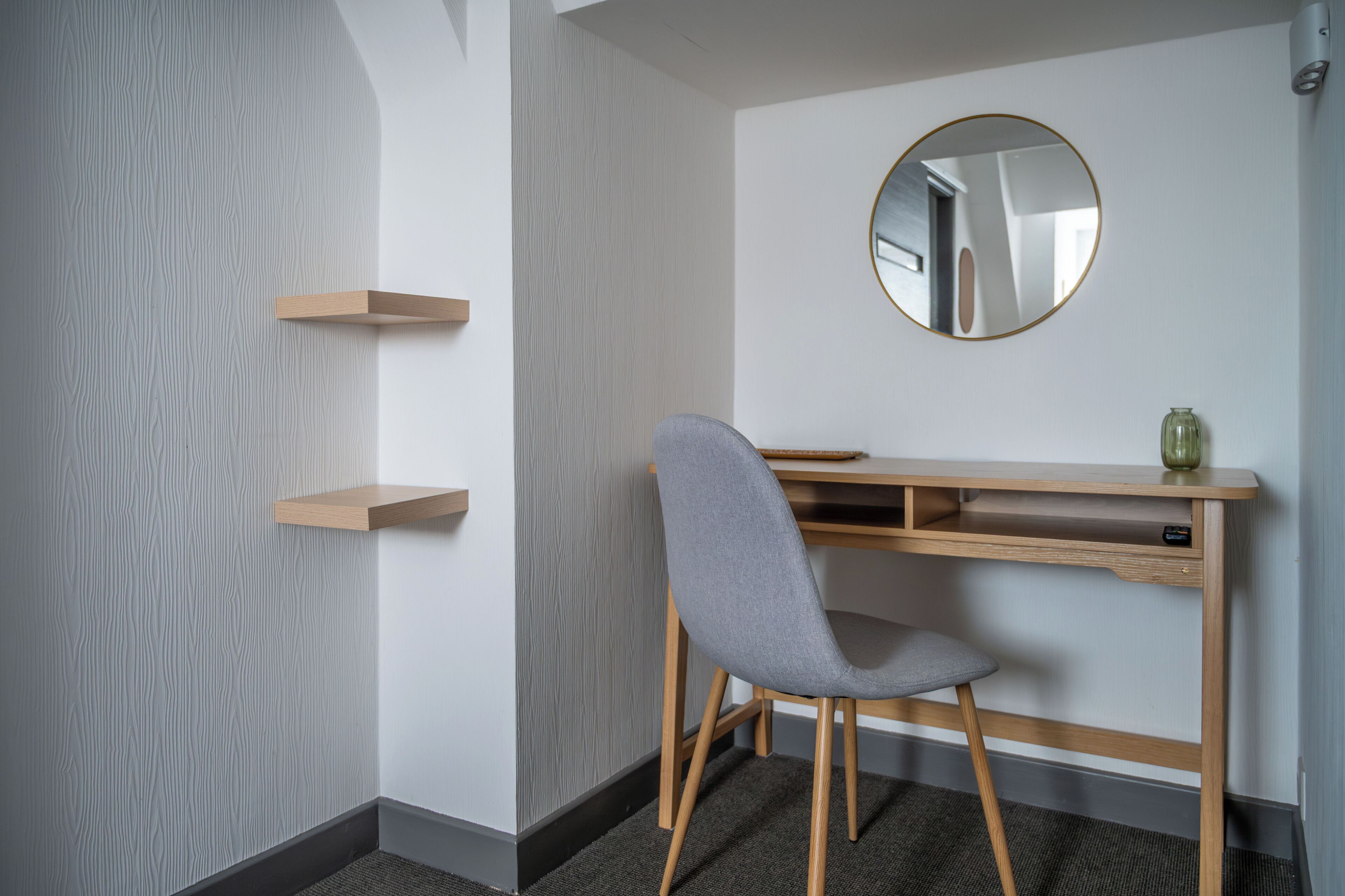Une chambre moderne avec un bureau en bois, une chaise grise, un miroir rond et deux étagères en bois.