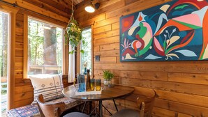 A wooden interior with a round table, chairs, a bench, a hanging plant, and a colorful abstract painting. - Barrel sauna, cedar cabin, woodstove (Deming)