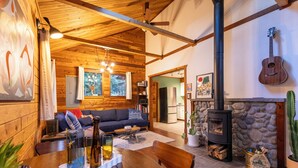 A cozy cabin interior with a wooden ceiling, a stone fireplace, a dining area, and a living room with a sofa and a guitar on the wall. - Barrel sauna, cedar cabin, woodstove (Deming)