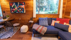 A cozy cabin interior with a wooden wall, a sofa with cushions, a table with chairs, and a window with a forest view. - Barrel sauna, cedar cabin, woodstove (Deming)