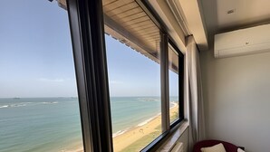 Ein Zimmer mit großem Fenster und Blick auf einen Strand, einem Heizkörper und einem Sessel mit Kissen.