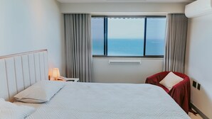 Ein Hotelzimmer mit Bett, Nachttisch, Lampe, Fenster mit Meerblick und einem roten Sessel.