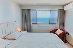Ein Hotelzimmer mit Bett, Nachttisch, Lampe, Fenster mit Meerblick und einem roten Sessel.