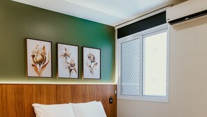 Ein ordentlich bezogenes Bett mit grüner Decke, weißen Kissen und gefalteten Handtüchern auf einer weißen Bettdecke. An der Wand über dem Bett hängen drei gerahmte Blumenbilder.