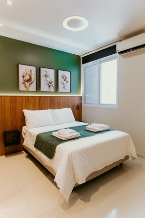Ein ordentlich bezogenes Bett mit grüner Decke, weißen Kissen und gefalteten Handtüchern auf einer weißen Bettdecke. An der Wand über dem Bett hängen drei gerahmte Blumenbilder.