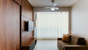 Ein modernes Wohnzimmer mit Flachbildfernseher, Deckenventilator, Sofa und einem großen Fenster mit Jalousien.