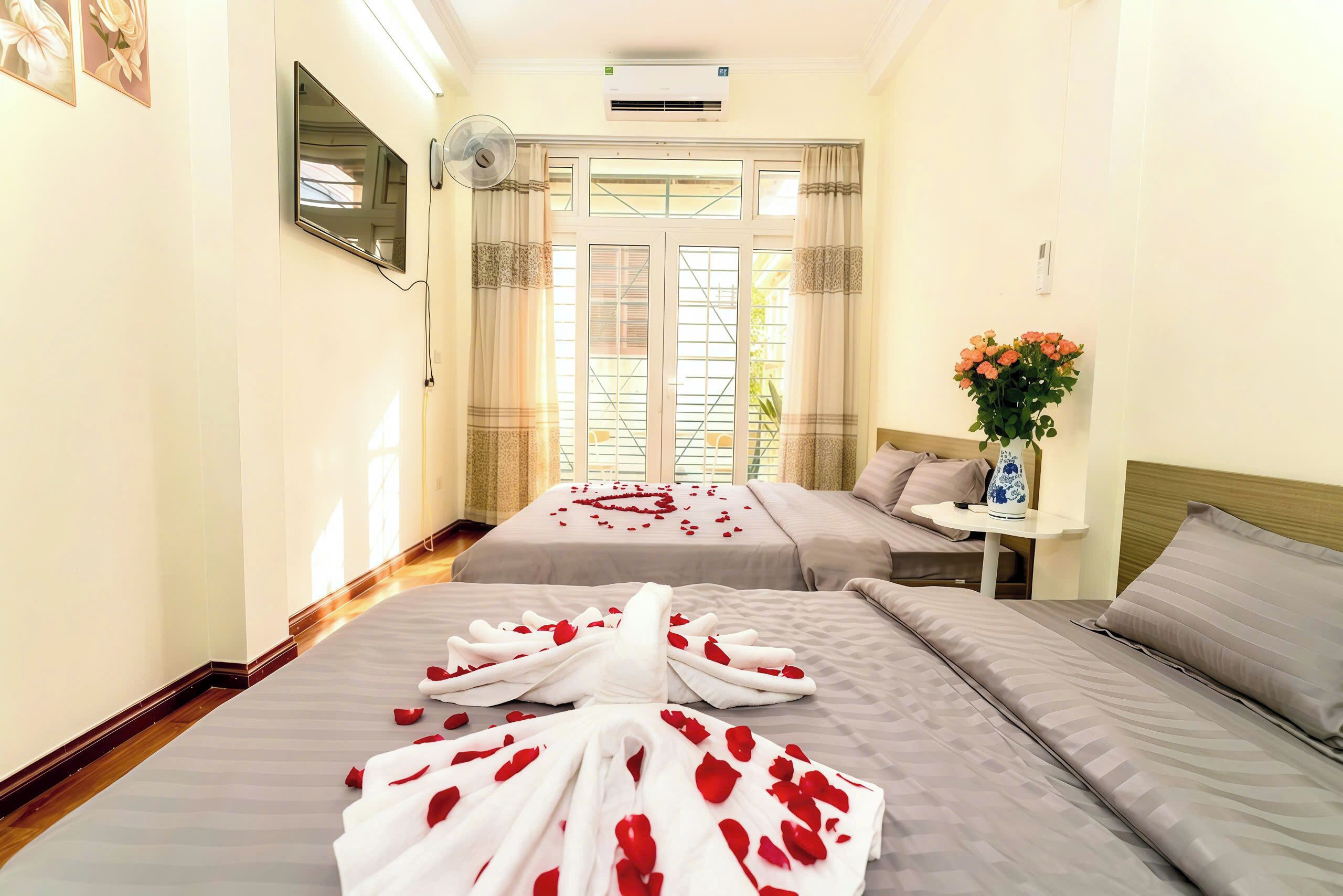 A hotel room with two beds, a TV, an air conditioner, a small table with a vase, and a window with curtains.