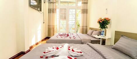 A hotel room with two beds, a TV, an air conditioner, a small table with a vase, and a window with curtains.