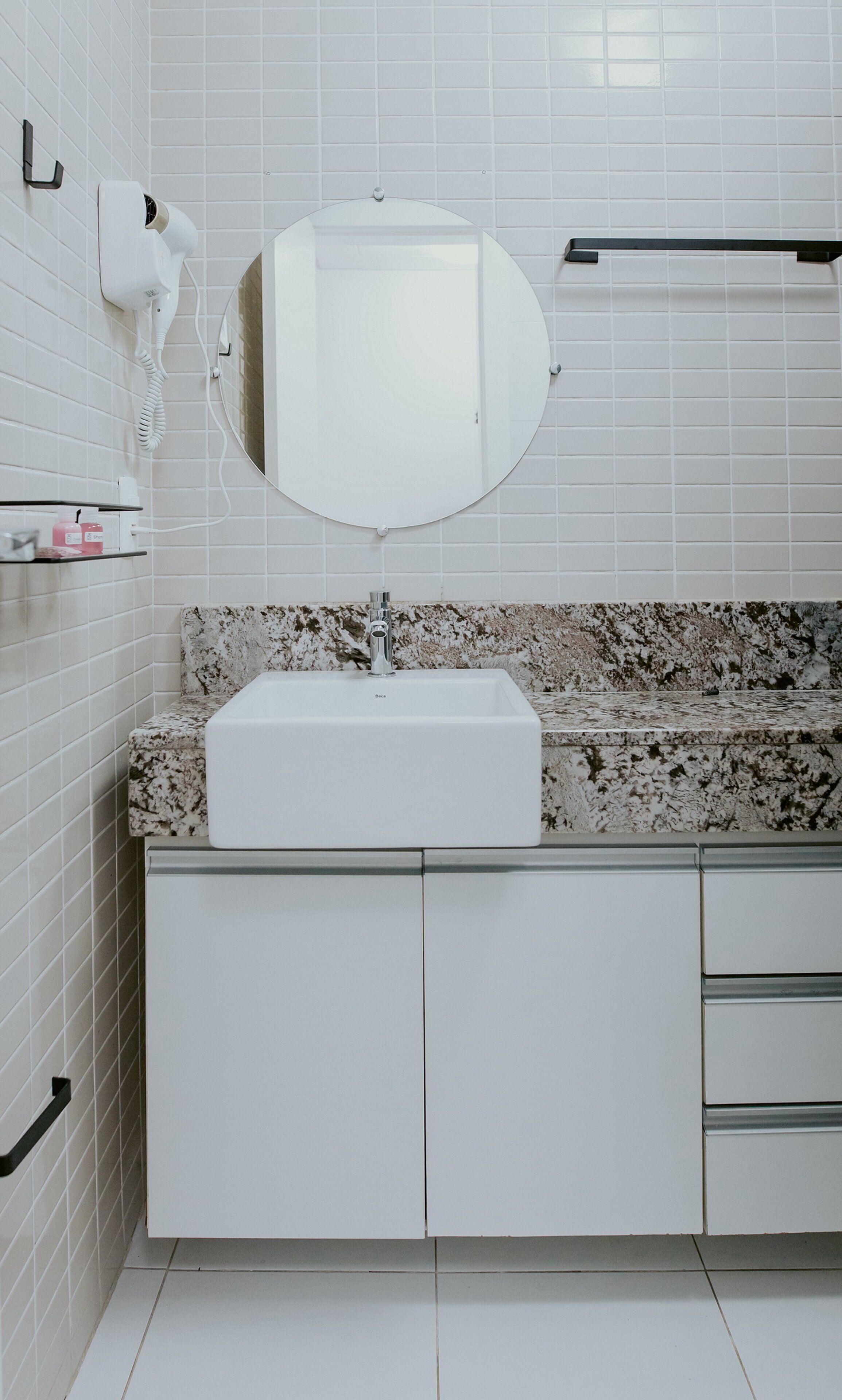Een badkamer met witte betegelde wanden, een granieten aanrecht, een witte wasbak, een spiegel en een handdoekhouder.