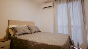 Un dormitorio con cama, una mesita de noche, una ventana con cortinas y un equipo de aire acondicionado.