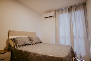 Un dormitori amb un llit, una tauleta de nit, una finestra amb cortines i un aparell d'aire condicionat.