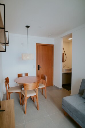 Uma sala de jantar com uma mesa redonda e cadeiras, uma porta de madeira e uma cozinha compacta ao fundo.