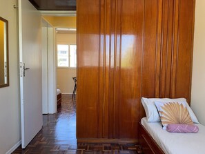 Ein Zimmer mit einem Holzschrank, einem Bett mit gemustertem Kopfkissen und einem Stuhl im Hintergrund.
