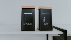 Twee zwarte containers met houten deksels, gelabeld met 'AÇÚCAR' en 'CAFÉ', staan op een witte plank.