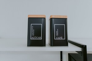 Zwei schwarze Behälter mit Holideckeln, beschriftet mit 'AÇÚCAR' und 'CAFÉ', auf einem weißen Regal.
