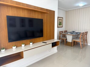 Ein modernes Wohnzimmer mit einem großen Flachbildfernseher an einer Holzwand, einem Essbereich mit Tisch und Stühlen sowie einer kleinen, weißen Kommode mit Zierpflanzen.