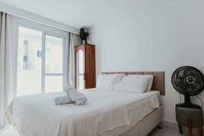 A hotel room with a bed, a wooden nightstand, a standing fan, and a window with curtains. - 2-bedroom apartment with the best location in Guarapari. (Guarapari)